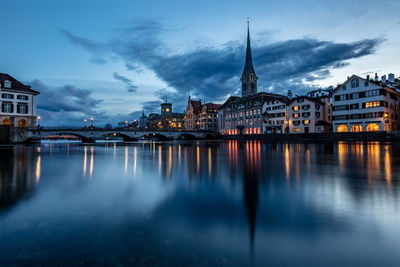 Illuminated buildings by river against cloudy sky