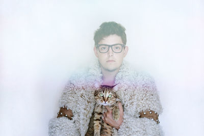 Portrait of young man wearing fur coat while holding cat against white background