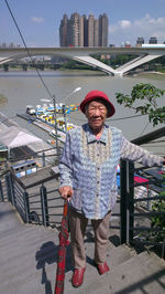 Portrait of senior woman with umbrella standing on steps against bridge over river