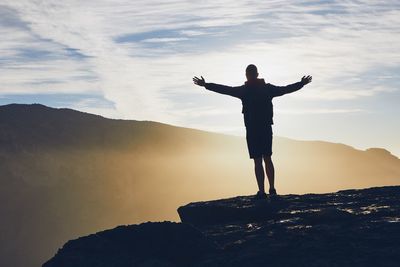 Man with arms outstretched standing at cliff against sky during sunset
