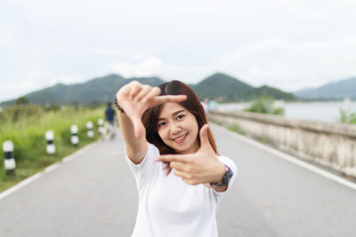 Portrait of smiling young woman making finger frame while standing on road