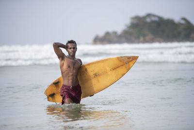 Portrait of shirtless man with surfboard wading in sea against sky