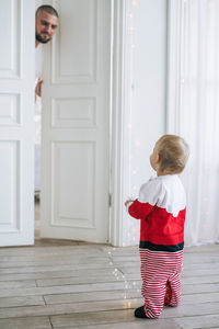 Cute little baby girl in santa costume standing in the large bright room looking at dad