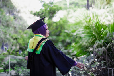 Rear view of asian woman wearing graduation gown standing against trees