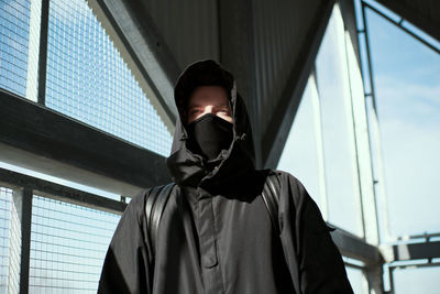 Thoughtful man wearing mask and hooded shirt standing by metallic grate