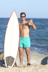 Portrait of shirtless young man with surfboard standing at beach against clear blue sky during sunny day