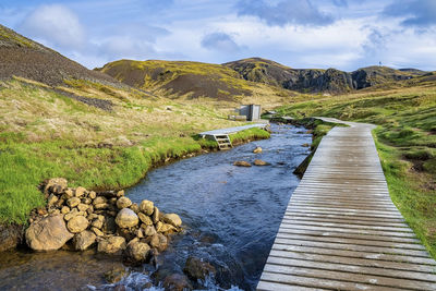 Wooden boardwalk by geothermal hot river stream in reykjadalur valley against sky