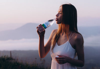 Young woman drinking water from bottle while standing against sky