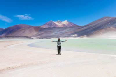 Young man with arms outstretched standing at desert against blue sky during sunny day