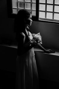 Thoughtful young woman with bouquet looking through window in darkroom