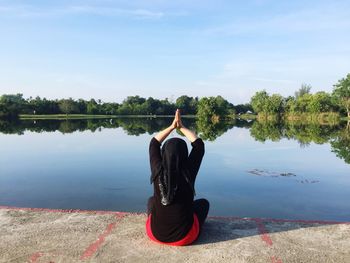Rear view of woman with hands clasped meditating while sitting by lake against sky