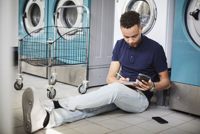 Young man holding mobile phone and studying while sitting at laundromat