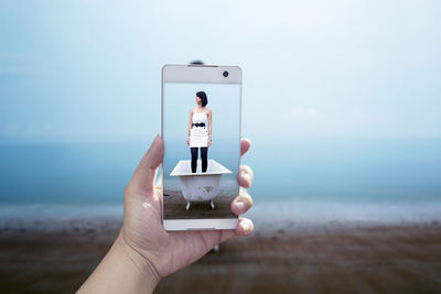 Cropped image of hand photographing woman standing in bathtub through mobile phone at beach