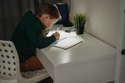 Caucasian boy doing homework at home in dark room by the light of lamp.