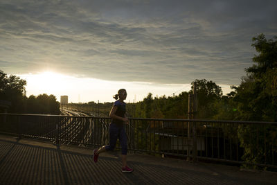 Woman running on road by railing against sky during sunset