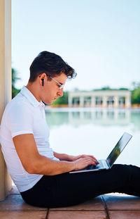 Side view of concentrated male student browsing netbook while sitting near stone column in university campus