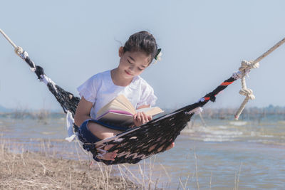 Girl reading book while sitting on hammock against sky