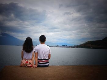 Rear view of couple sitting on pier by lake against cloudy sky