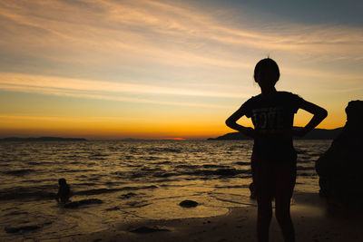 Girl standing on shore at beach against sky during sunset