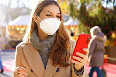 Beautiful young woman wearing medical face mask using smartphone on christmas time outdoors