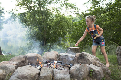 Blonde girl in overalls roasts marshmellow over campfire