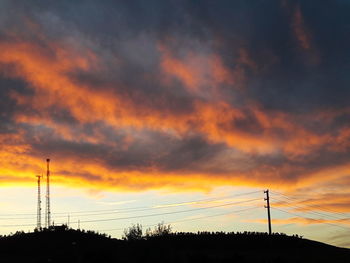 Low angle view of silhouette electricity pylons against orange sky