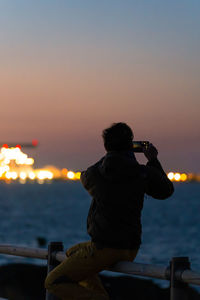 Rear view of man photographing sea against sky during sunset