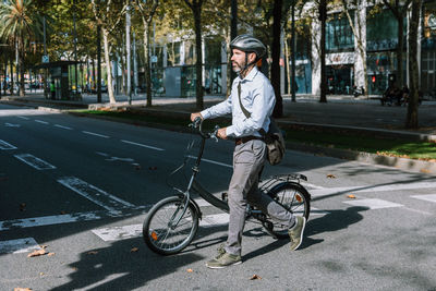 Full length of bearded male worker in helmet crossing asphalt roadway with bicycle on way to work