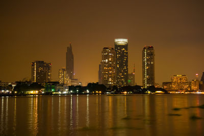 Illuminated buildings by river against sky at night