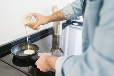 Man cooking in the kitchen in a denim shirt. an anonymous man is pouring a crepe mix into the pan
