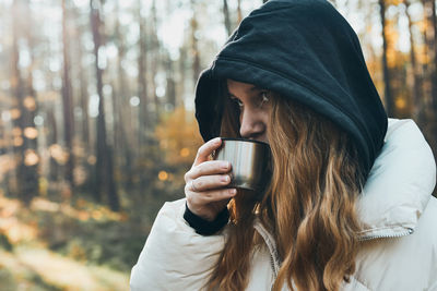 Woman in a hood having break during autumn trip holding cup with hot drink from thermos flask