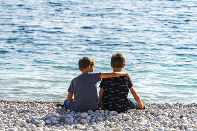 Two boys sitting on the beach, talking and throwing rocks into the sea