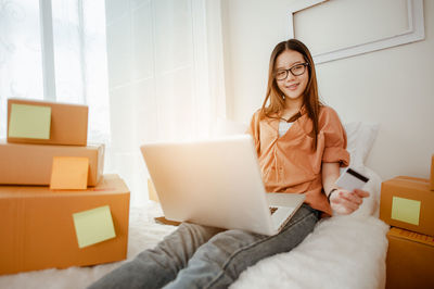 Smiling young woman holding credit card using laptop on bed in home office