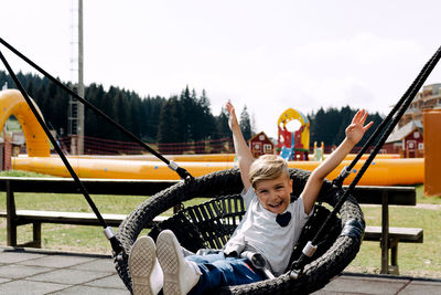 Cheerful kid with raised arms having fun while swinging at the playground.