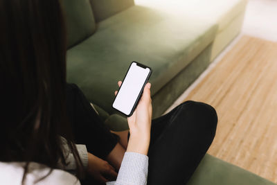 Black hear girl sitting on green sofa and holding smartphone in her hand with empty white screen