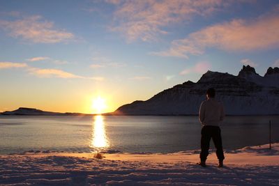 Rear view of man standing by lake against sky during sunset