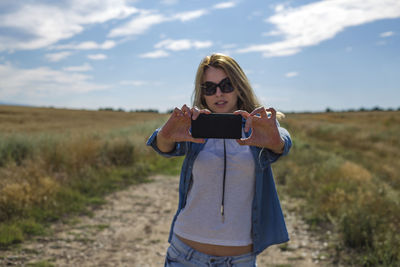 Mid adult woman taking selfie while standing on field against sky during sunny day