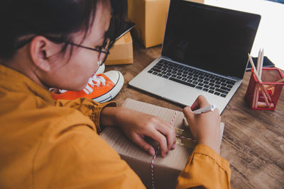 High angle view of girl writing over box by laptop on table