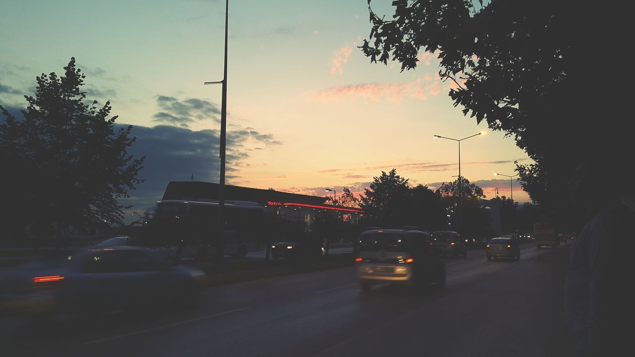 transportation, car, land vehicle, mode of transport, road, street, tree, sky, sunset, building exterior, city, on the move, traffic, the way forward, city street, architecture, street light, built structure, illuminated, silhouette