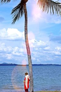 Woman standing by palm tree by sea against sky