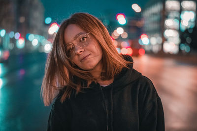 Portrait of young woman standing by illuminated street at night