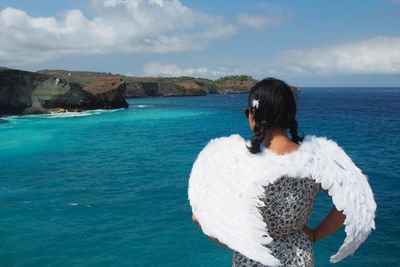 Rear view of woman with white costume wings looking at sea against sky
