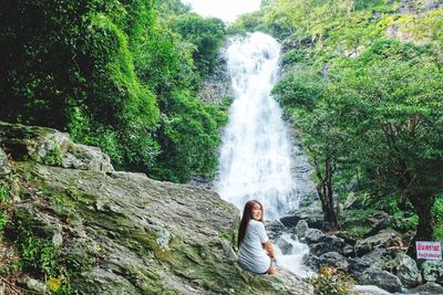 Rear view portrait of smiling woman sitting by waterfall in forest