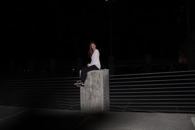 Full length of young woman sitting on concrete structure by railing at night