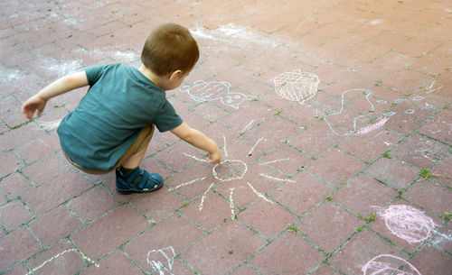 Little toddler boy two or three years old draws sun with crayons in the park on tiles.