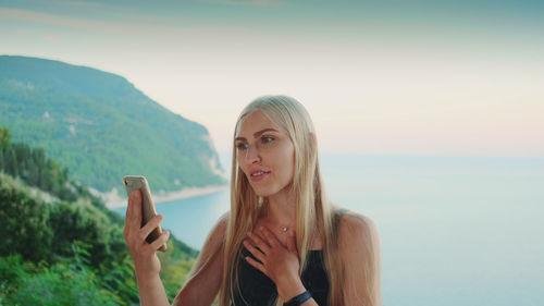 Portrait of young woman using phone by sea against sky