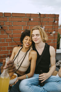 Smiling male and female friends sitting together with eyes closed during rooftop party