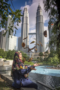 Happy woman wearing hijab throwing autumn leaves while sitting against petronas towers