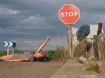 Man lying on road pointing at stop sign against cloudy sky
