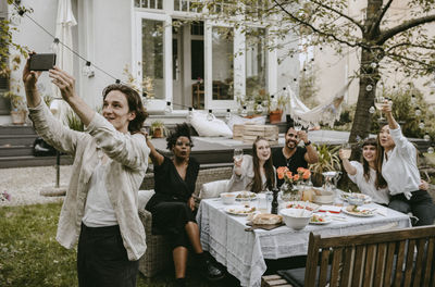 Smiling man taking selfie with friends through smart phone in yard during social gathering